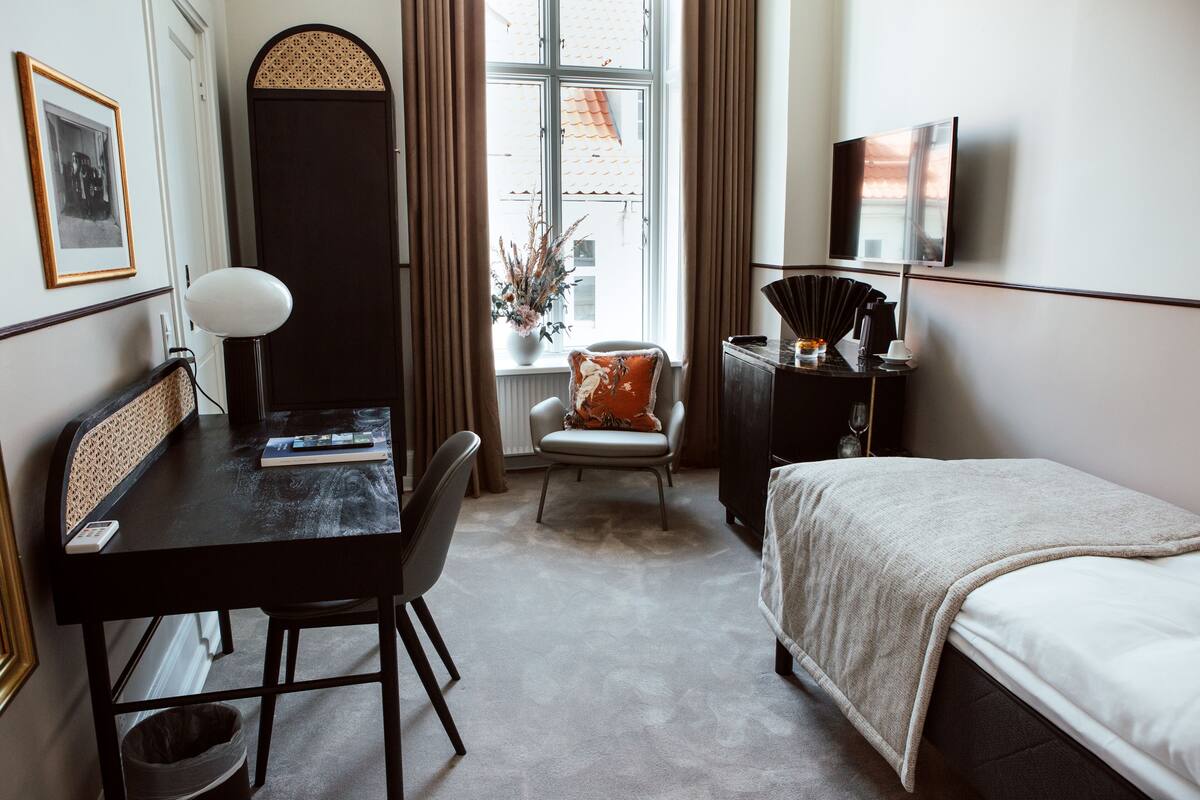 A hotel room with a bed, a desk with a chair, a TV, a window with curtains, and a small plant.