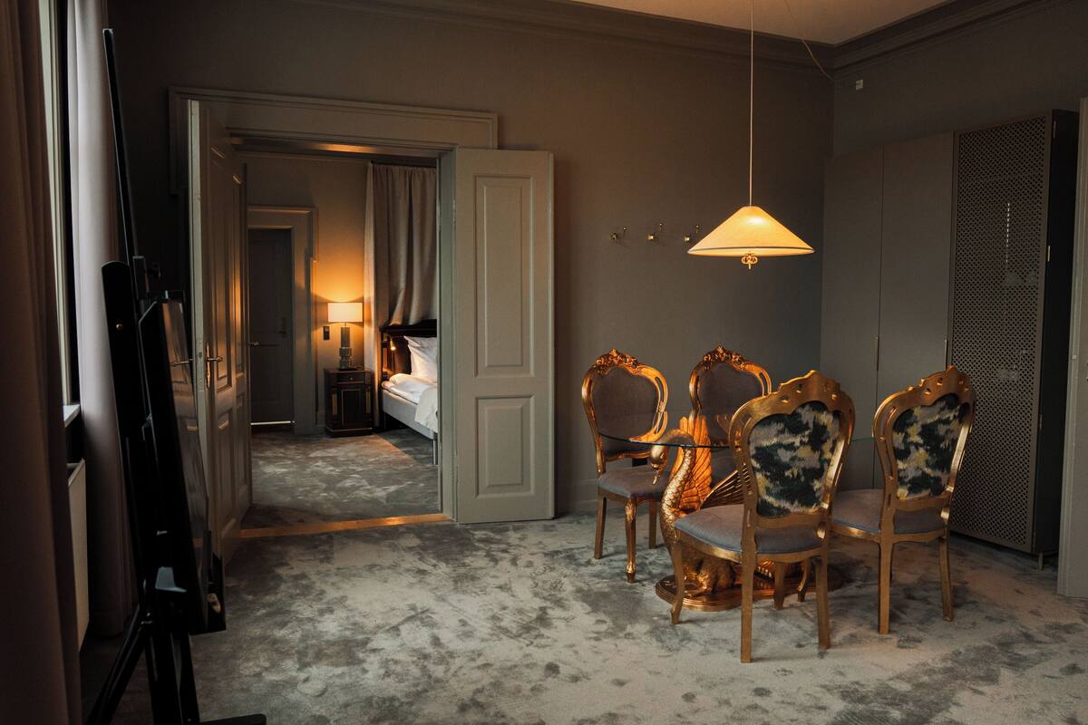 A hotel room with a bed, a desk, and a dining table with chairs. There is a lamp hanging from the ceiling.