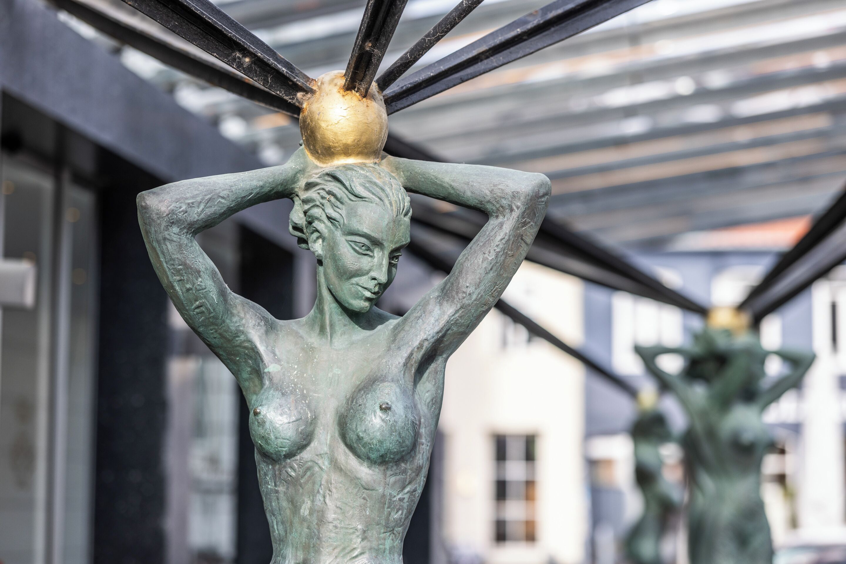 Escultura de una mujer con los brazos levantados y un tocado, ubicada al aire libre con edificios en el fondo.