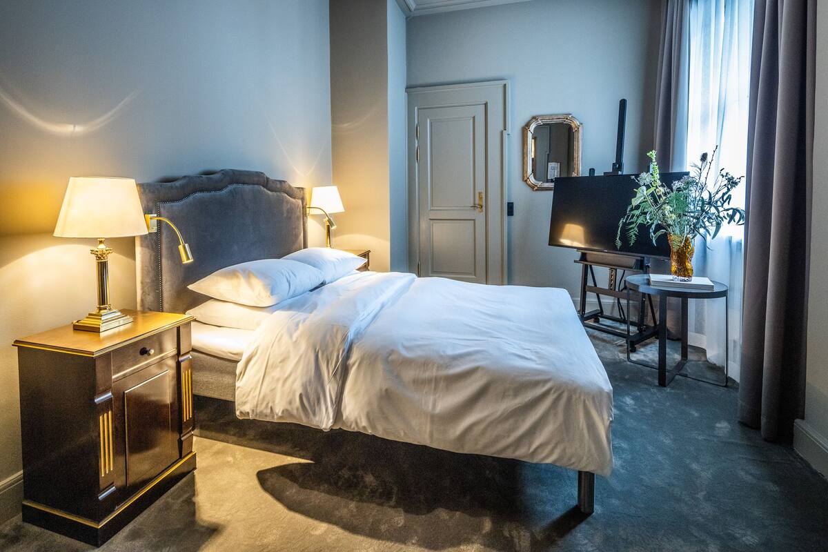 A hotel room with a bed, bedside lamps, a dresser, a desk with a chair, a mirror, and a potted plant.