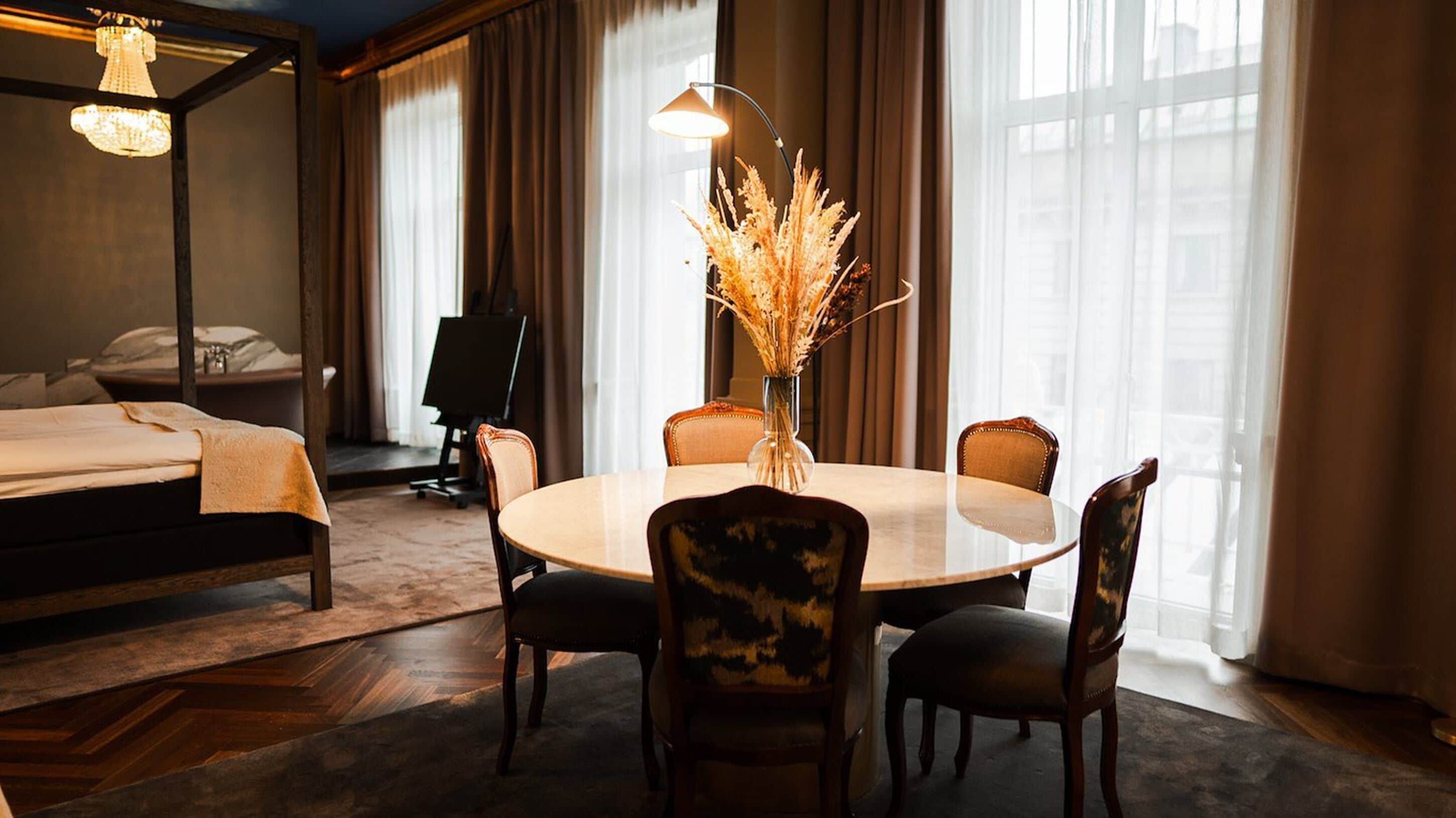A hotel room with a round dining table, four chairs, a vase with dried plants, a bed with a canopy, and a chandelier.