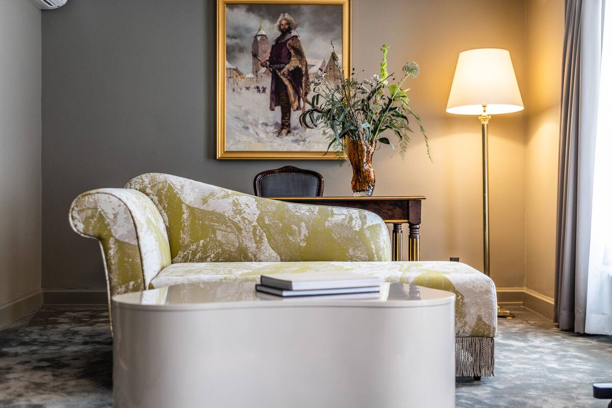 A room with a green and white patterned sofa, a white side table with a book, a vase with green plants, a painting on the wall, and a floor lamp.