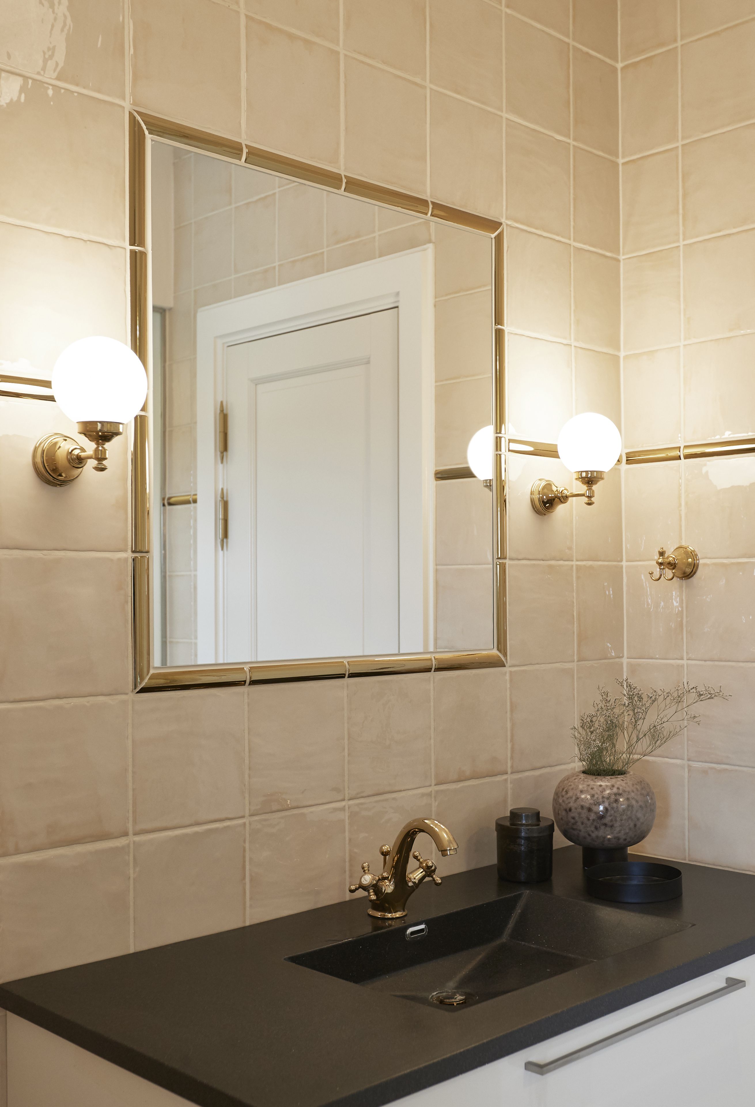 Een badkamer met een grote spiegel, twee wandlampen, een zwart wastafelmeubel en een vaas met gedroogde bloemen.