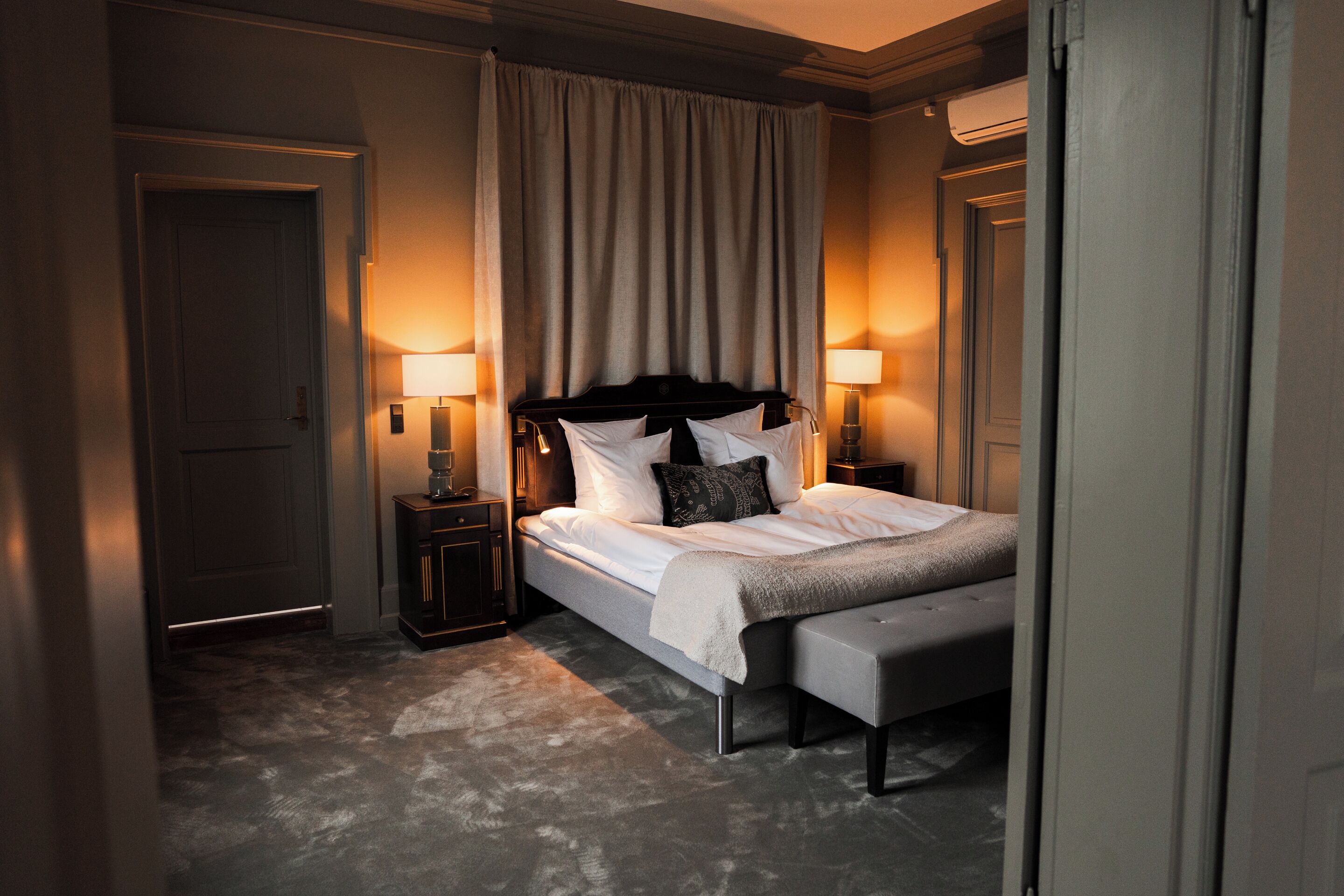 A hotel room with a bed, bedside lamps, a bench, and a door.