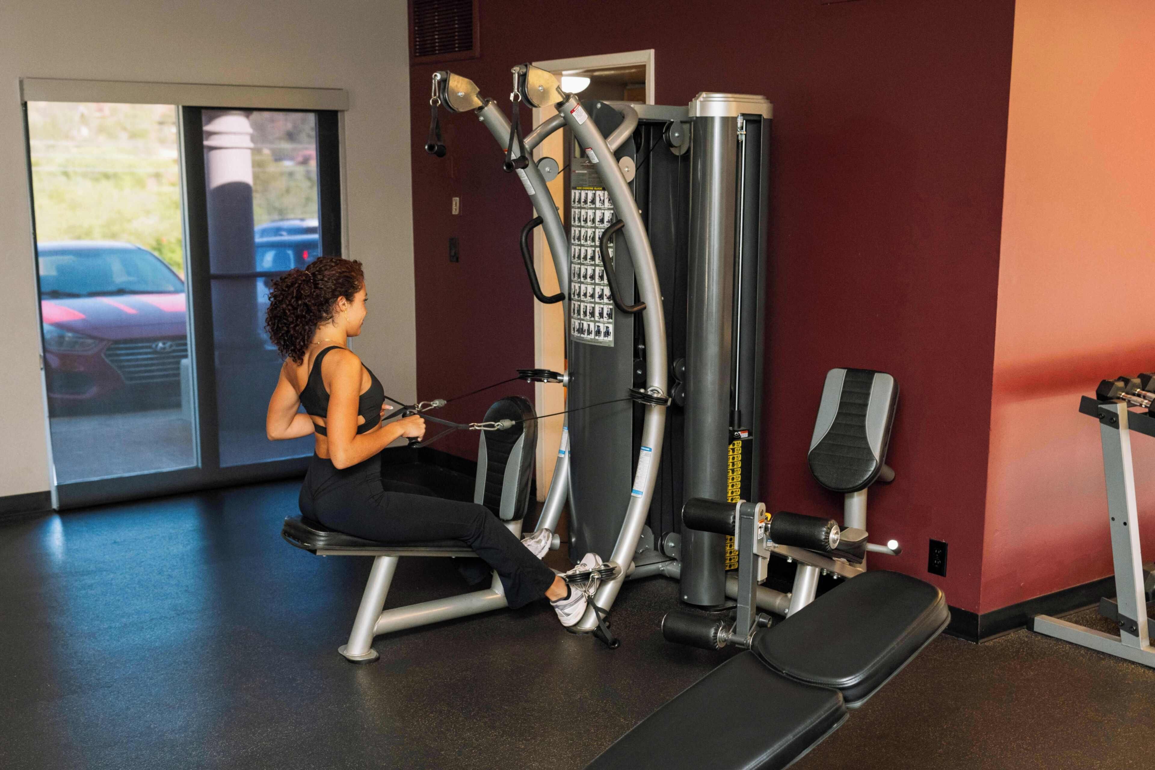A woman using a leg press machine in a gym with a bench and a wall-mounted exercise machine.
