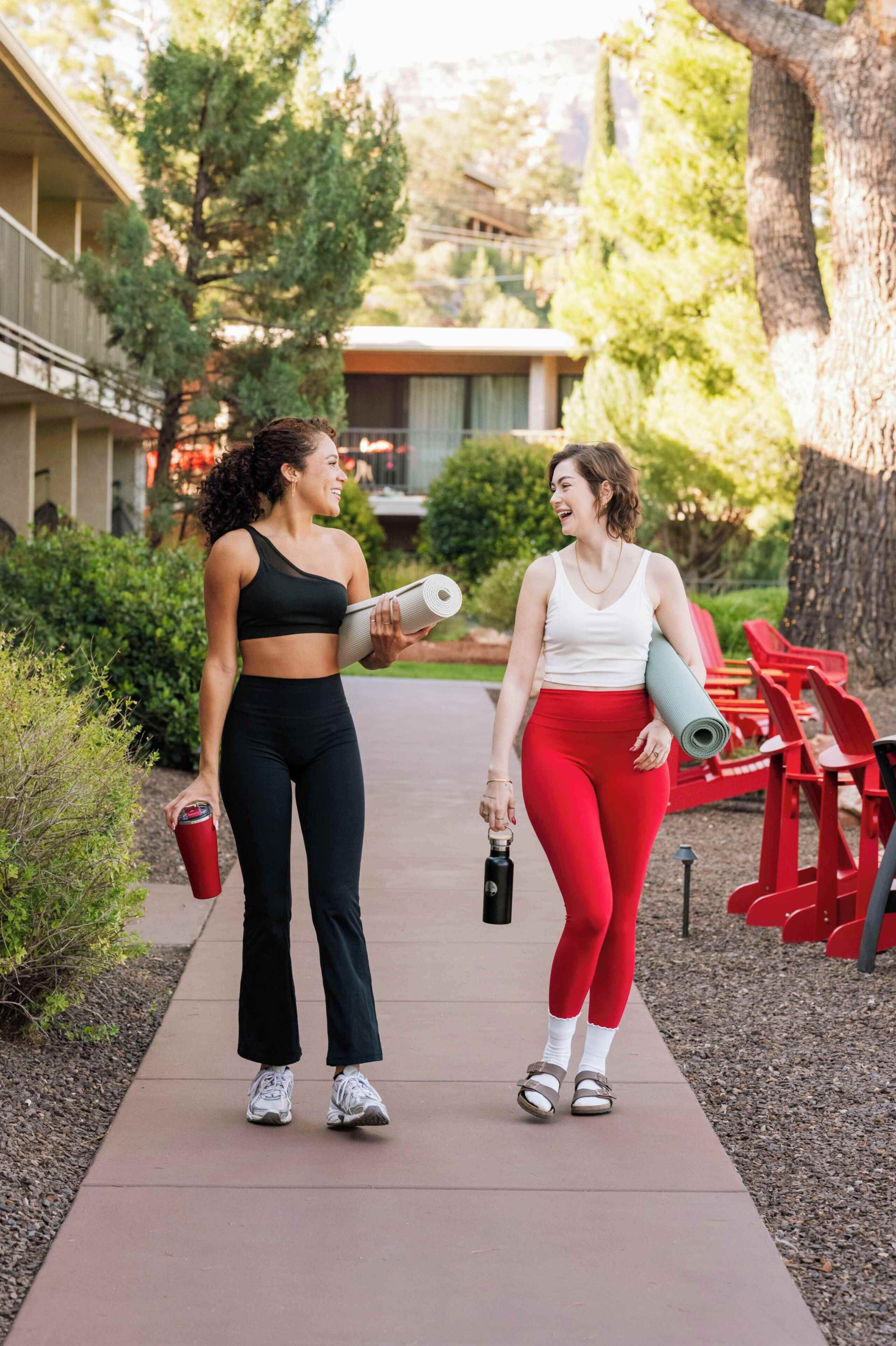 Dos personas caminando por un sendero con esterillas de yoga, una sosteniendo una botella de agua, y sillas rojas al fondo.