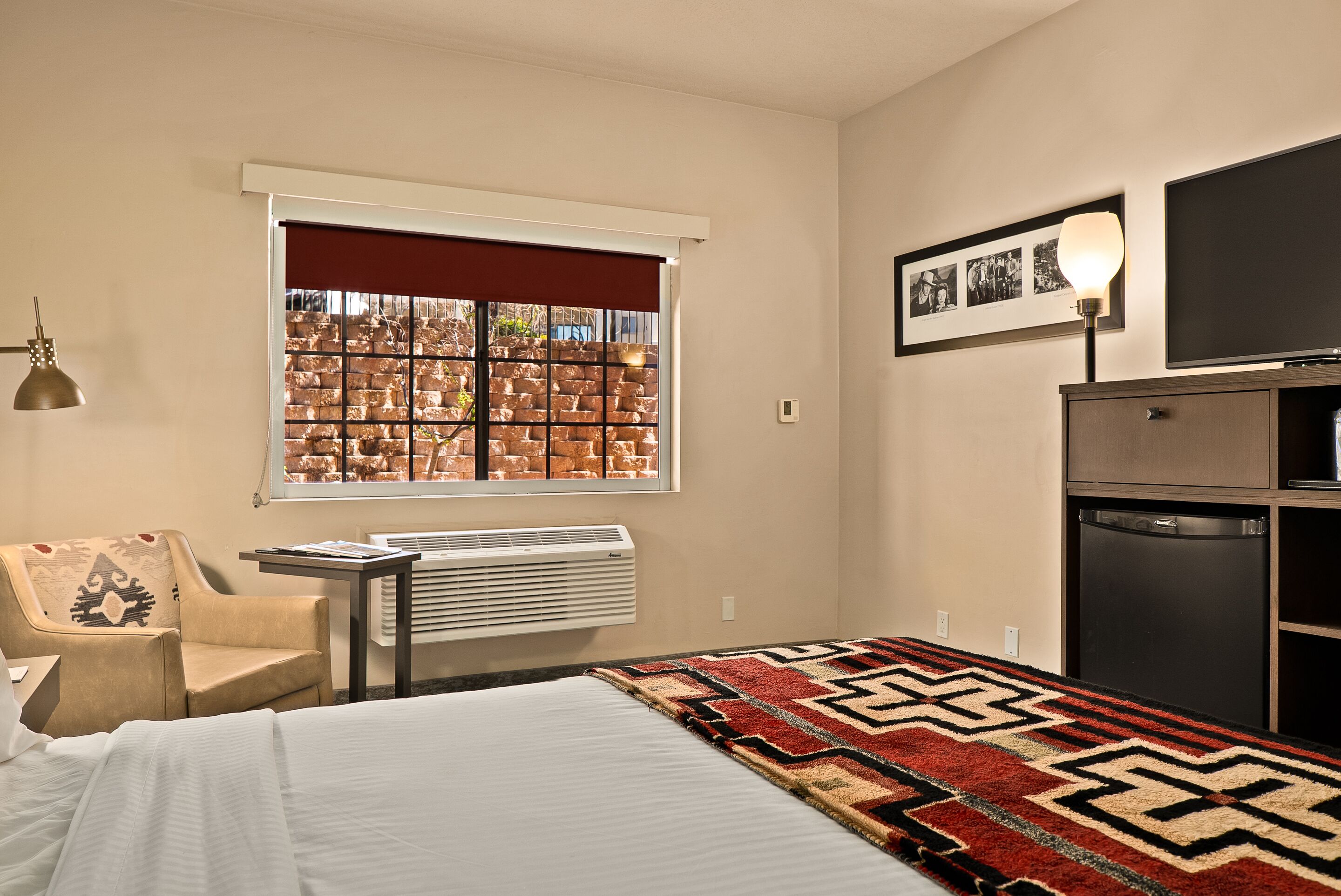 A hotel room with a bed, a chair, a window with a view of a brick wall, a radiator, a desk, and a television.