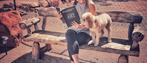 Une personne assise sur un banc en bois, en train de lire un livre, avec un petit chien debout à côté d’elle.