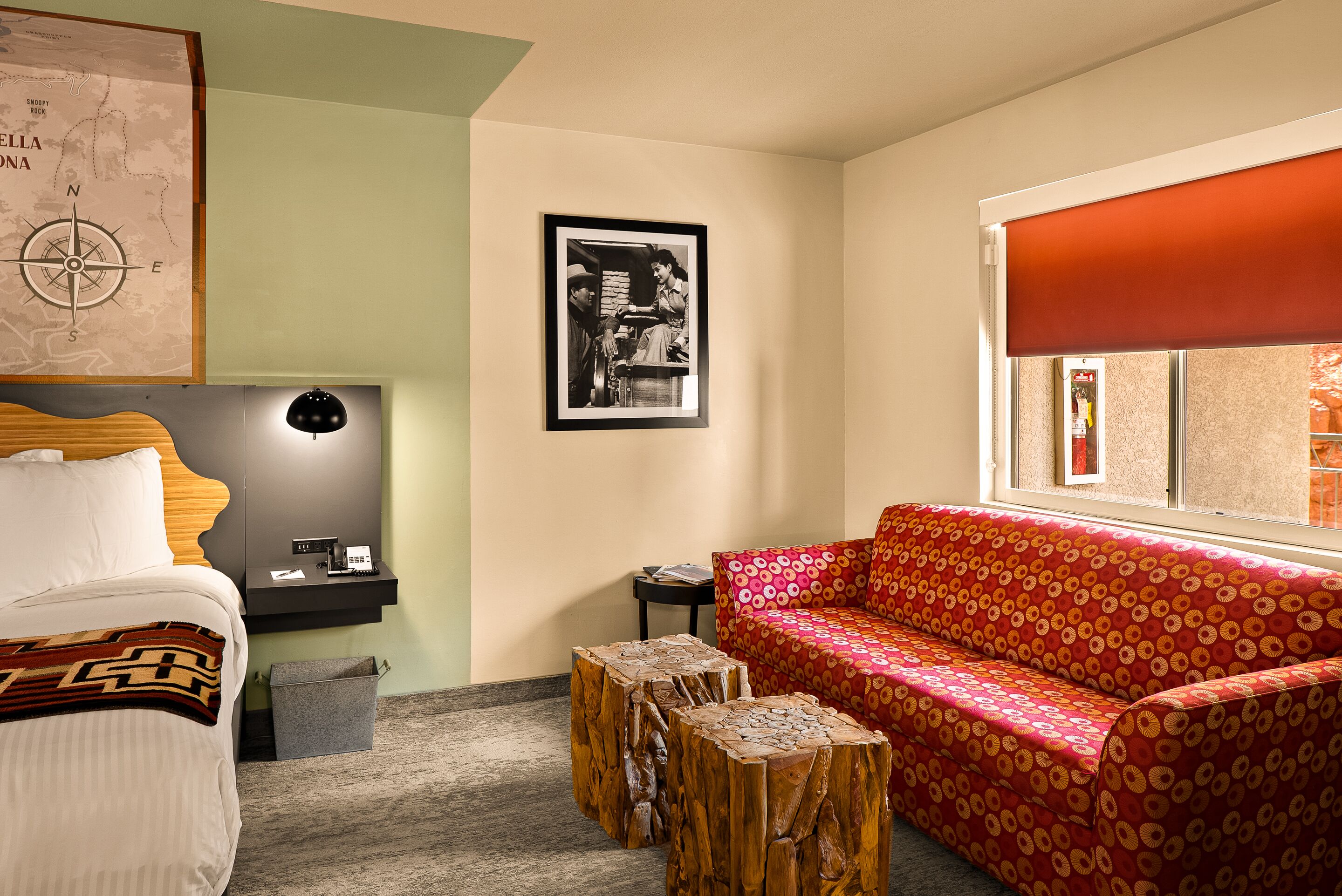 A hotel room with a bed, a red patterned sofa, a wooden coffee table, a map on the wall, and a framed picture.