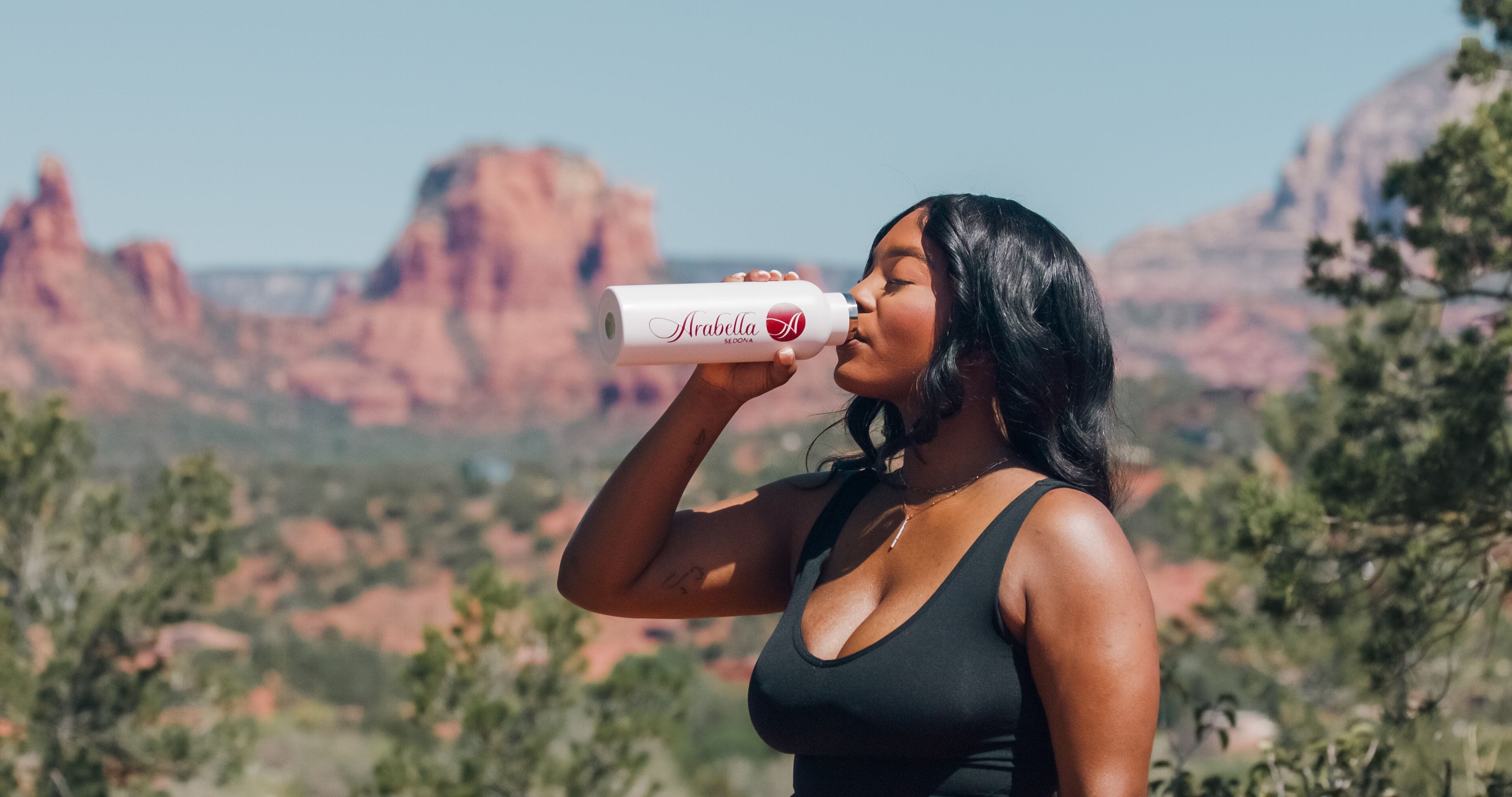 A person holding a bottle labeled "Arabella Sedona" outdoors with a mountainous landscape in the background.