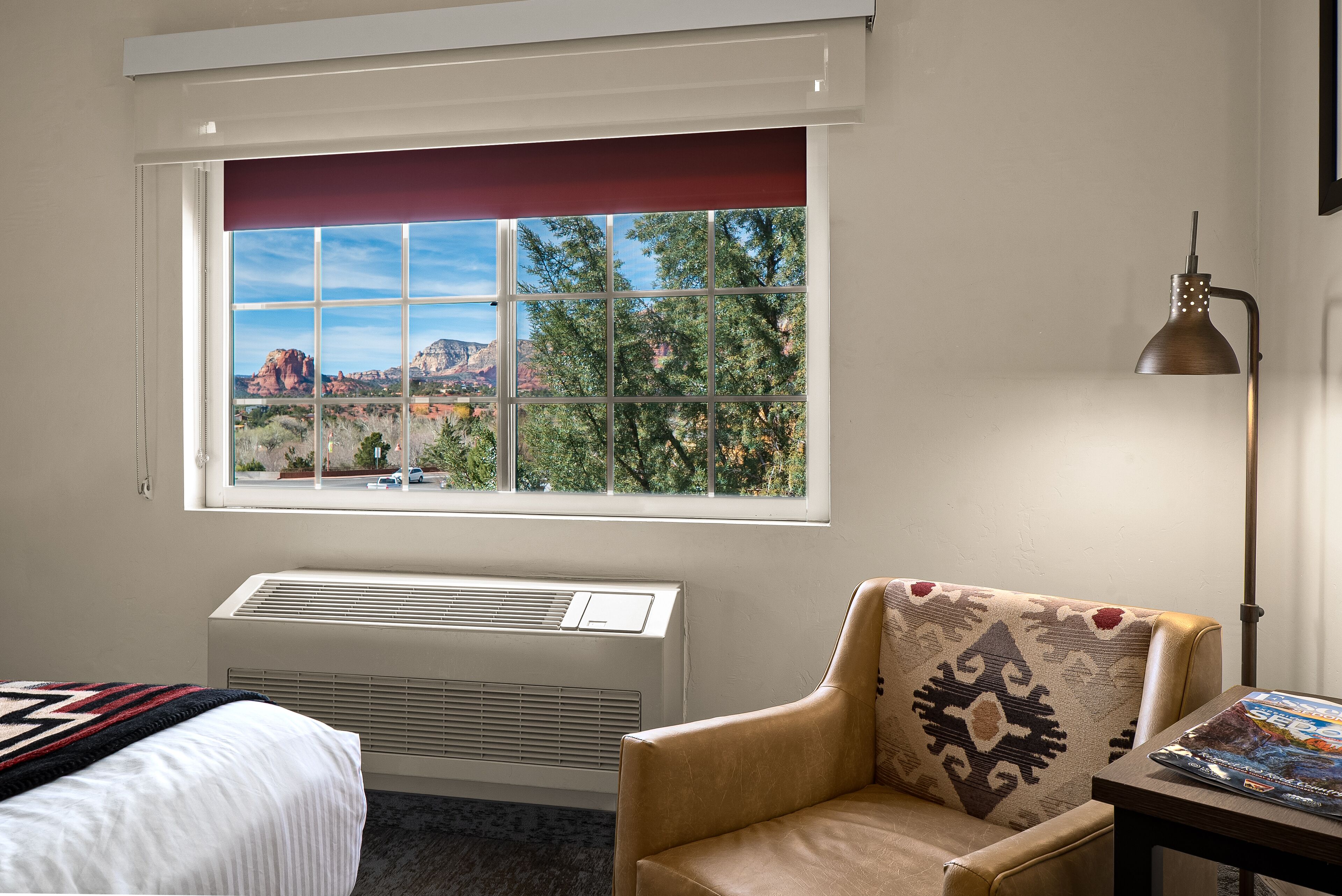 Ein Hotelzimmer mit großem Fenster, einem Bett, einem Sessel, einem Tisch und einer Lampe.