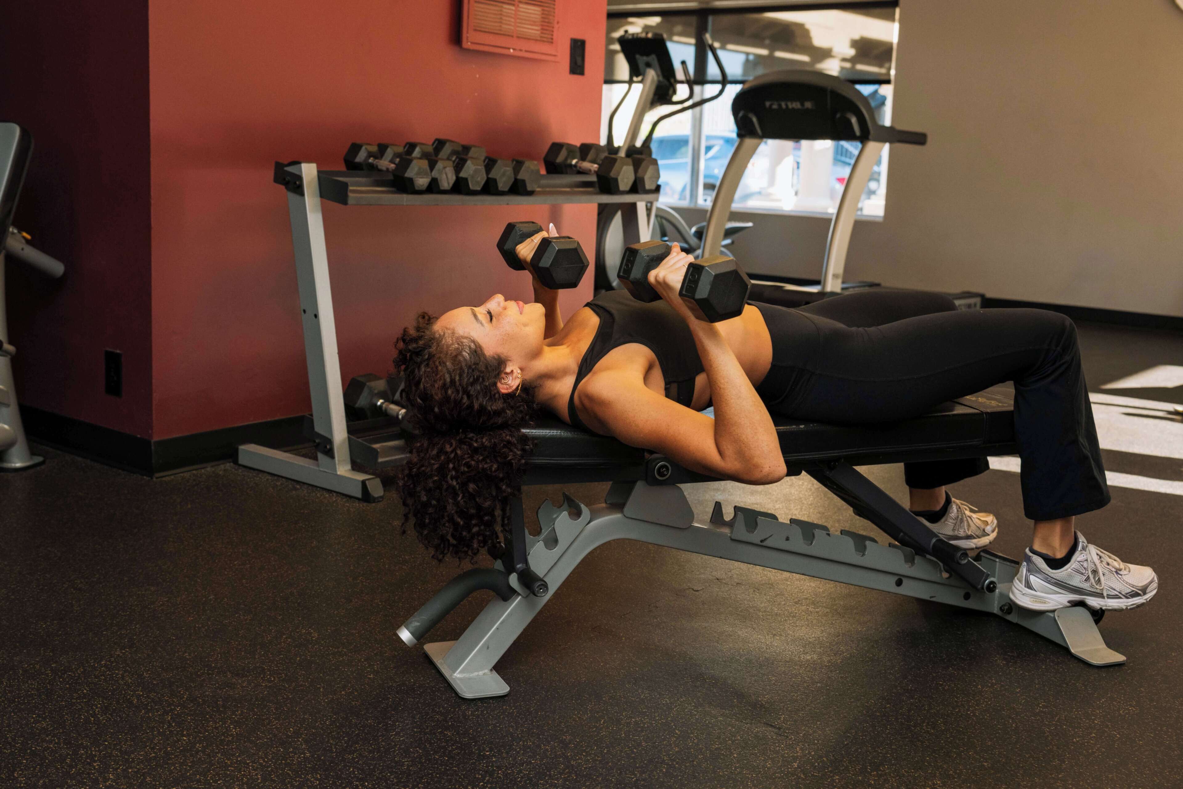 En person ligger på en bænk i et fitnesscenter og laver dumbbelløvelse.