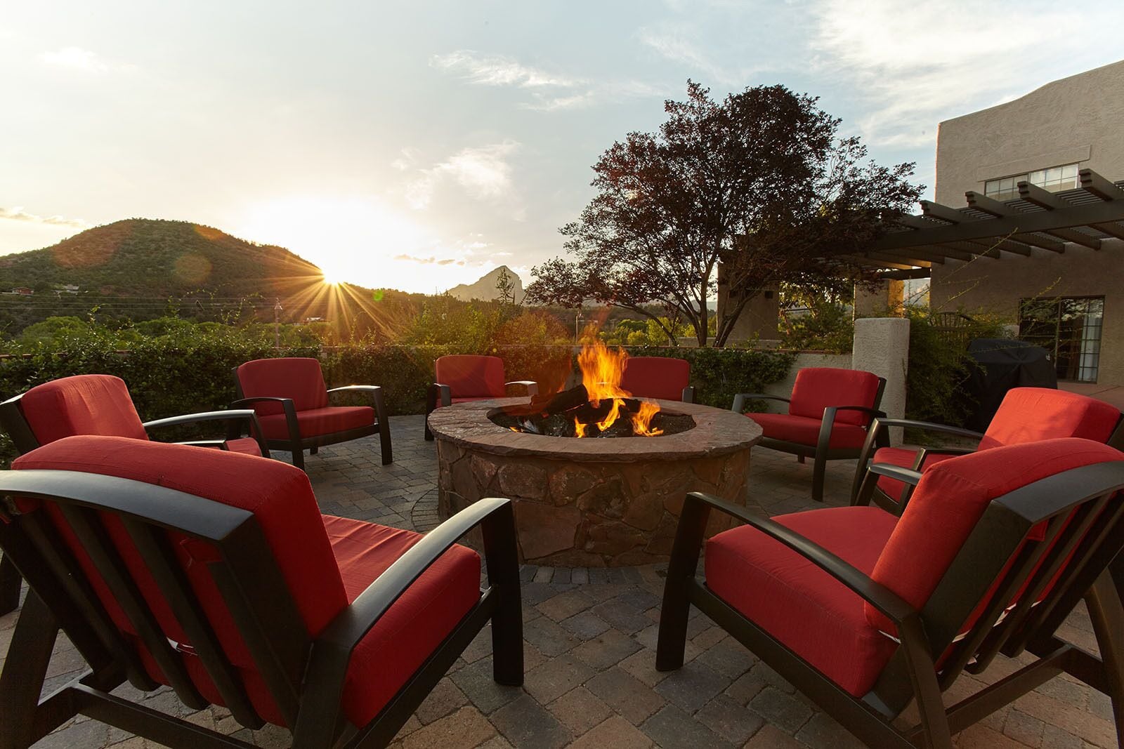 A patio with red chairs around a stone fire pit, set against a backdrop of hills and a building.