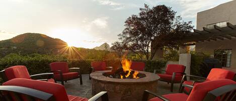 A patio with red chairs around a stone fire pit, set against a backdrop of hills and a building.