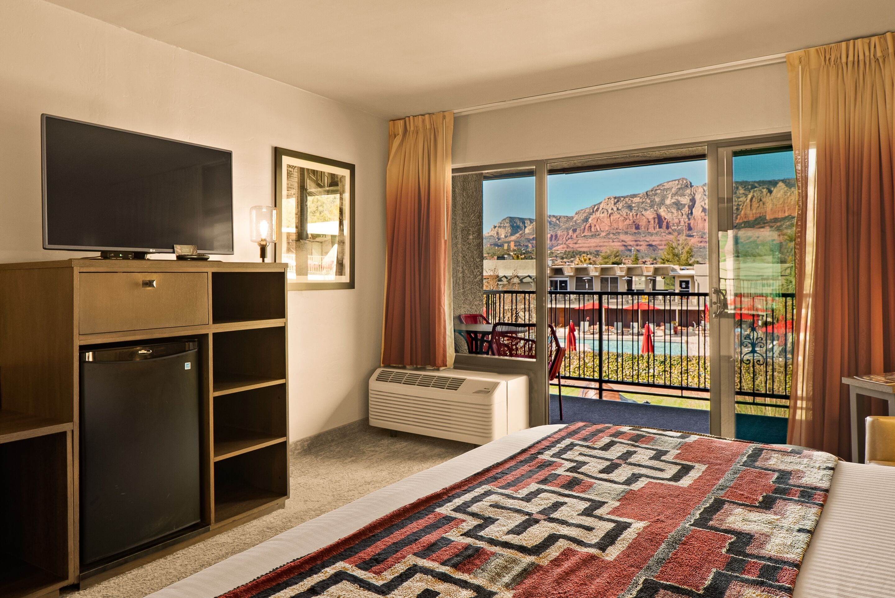 A hotel room with a bed, a TV, a mini-fridge, a heater, and a balcony with a view of mountains.