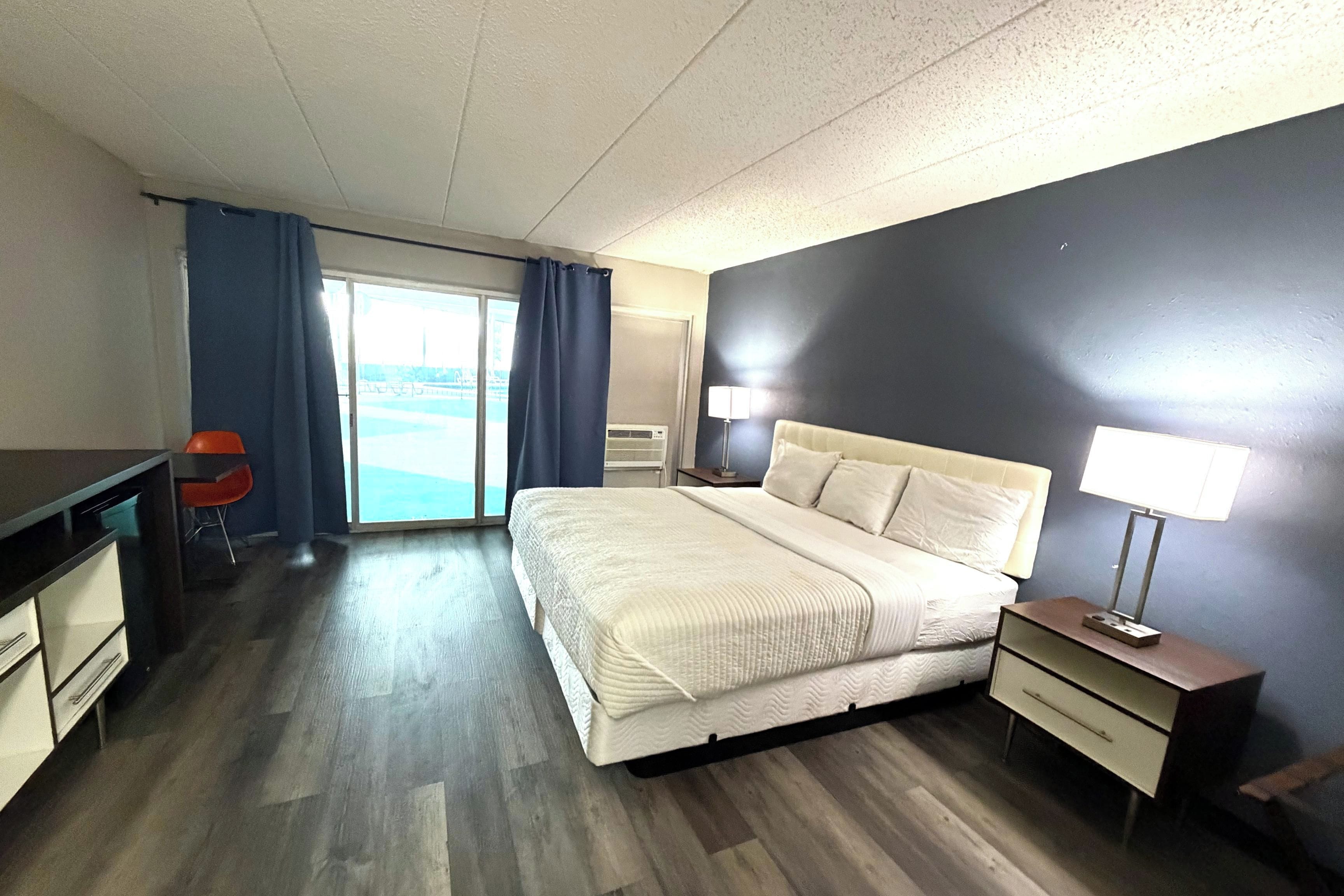A hotel room with a bed, bedside table, lamp, desk, chair, and a view of the outdoors.