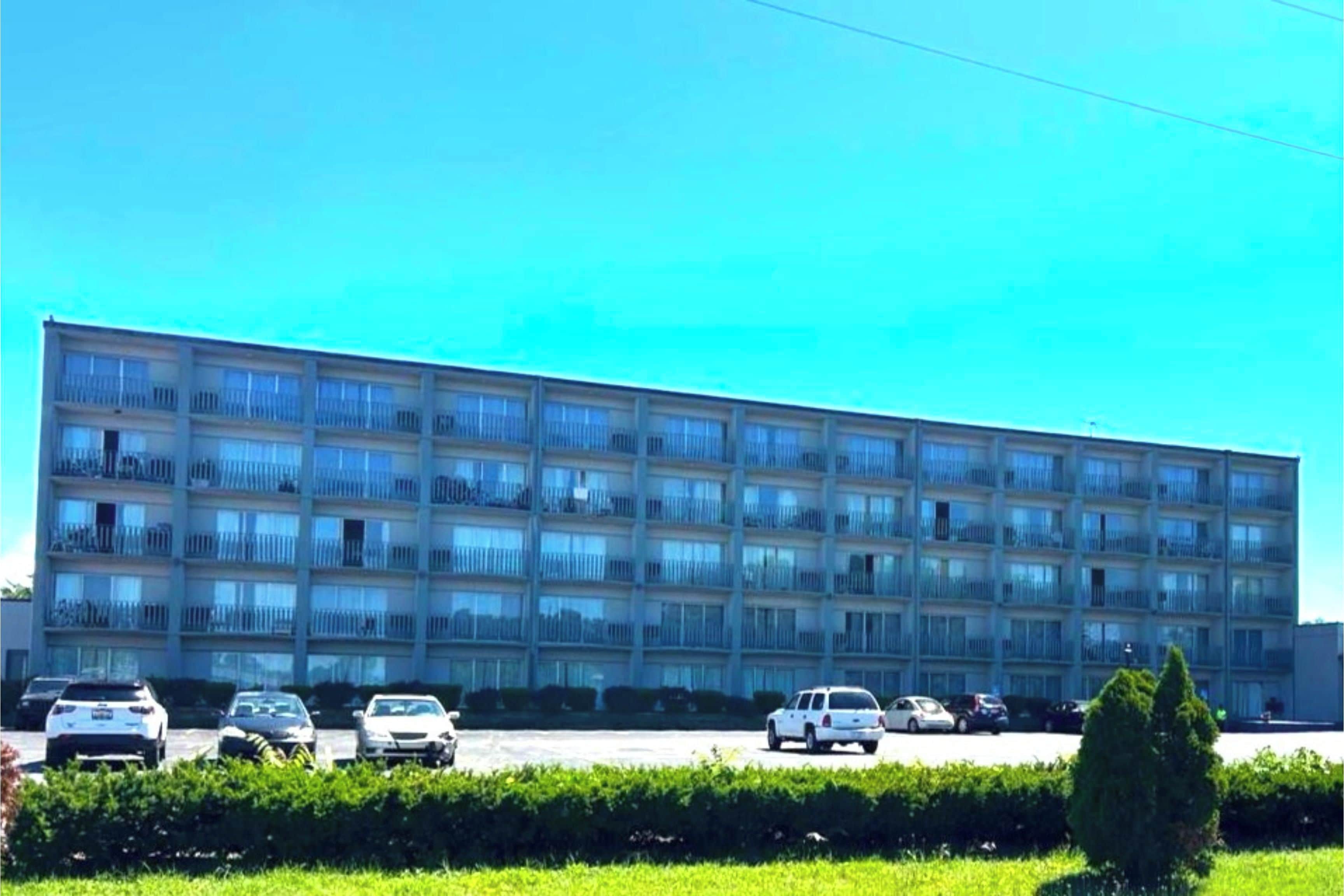Ein mehrgeschossiges Gebäude mit einer flachen Fassade, mehreren Balkonen und einem Parkplatz mit verschiedenen Autos.