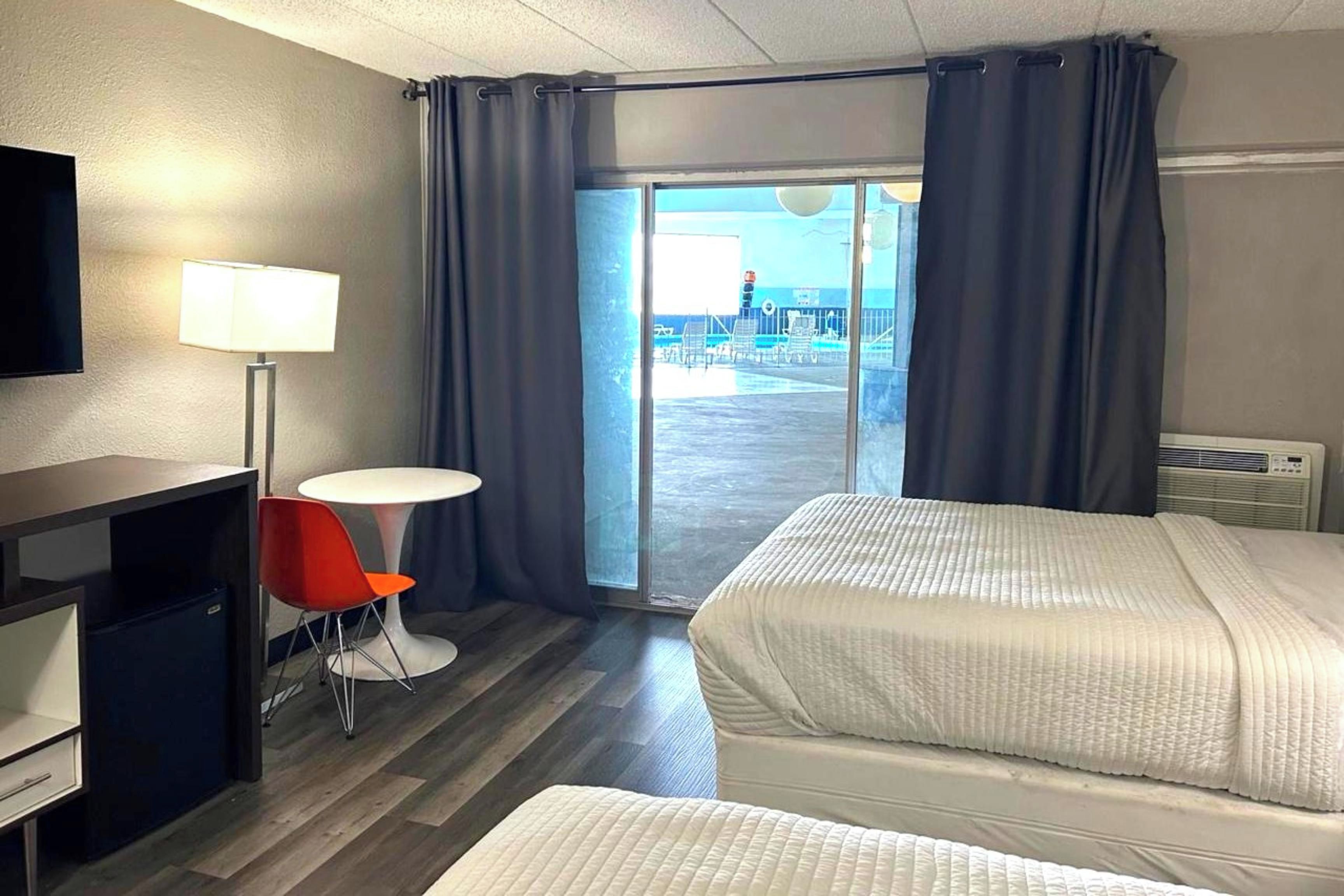 A hotel room with two beds, a desk, a chair, a lamp, and a view of the pool area.