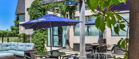 An outdoor seating area with a sofa, tables, and chairs, shaded by umbrellas, and a building with large windows in the background.