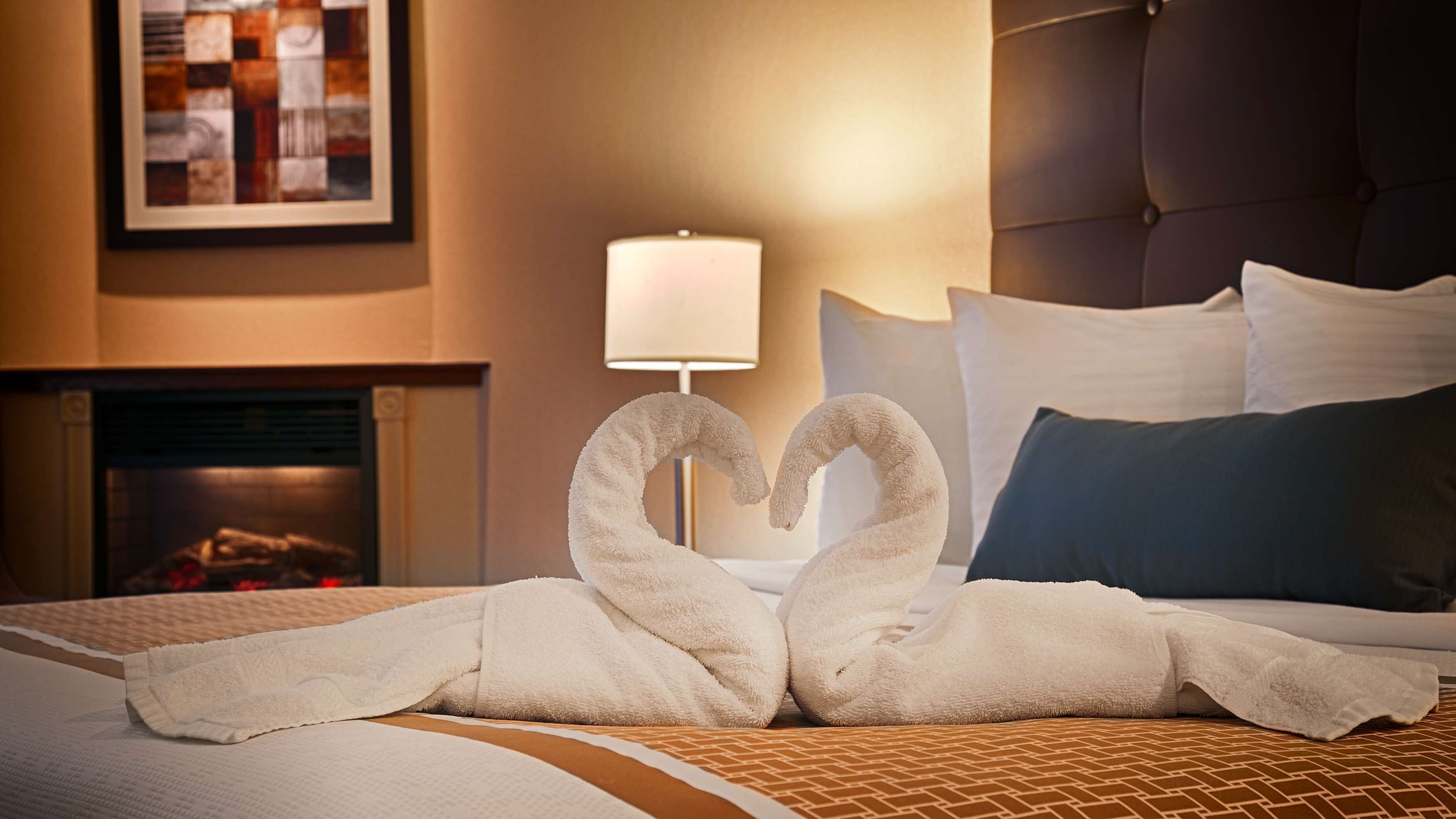 A neatly made bed with a heart-shaped towel arrangement, a bedside lamp, and a framed picture on the wall.
