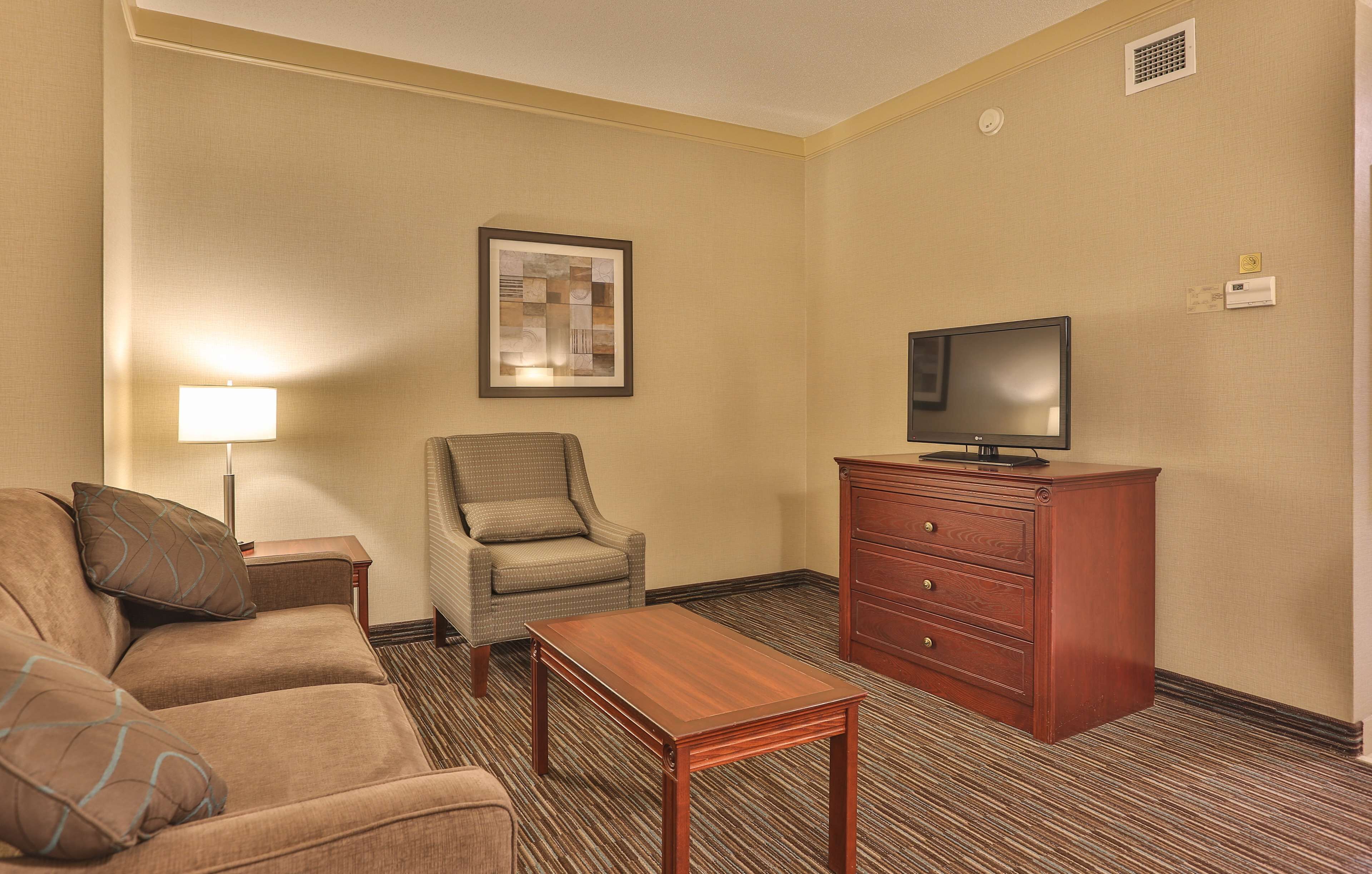 A hotel room with a sofa, a chair, a wooden table, a TV on a wooden cabinet, and a framed picture on the wall.