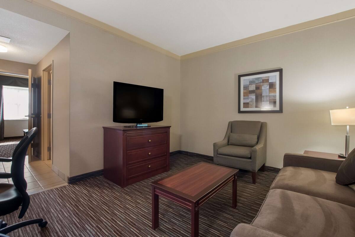 Habitación de hotel con televisor de pantalla plana sobre un mueble de madera, un sillón gris, un sofá marrón, una mesita de madera y un cuadro enmarcado en la pared.