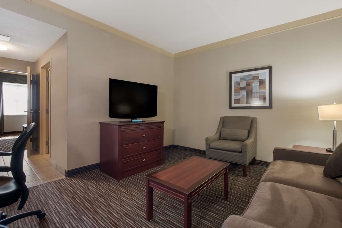 Habitación de hotel con televisor de pantalla plana sobre un mueble de madera, un sillón gris, un sofá marrón, una mesita de madera y un cuadro enmarcado en la pared.