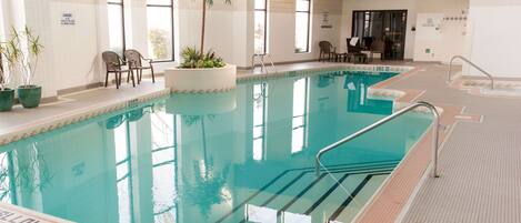 An indoor swimming pool with clear water, a safety sign, and a few chairs and potted plants around the pool area.