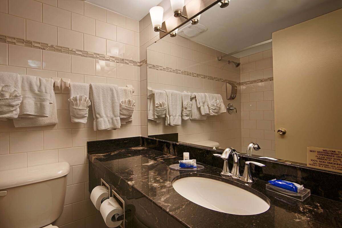 Un baño con inodoro, lavamanos y espejo. Se observan toalleros en la pared.