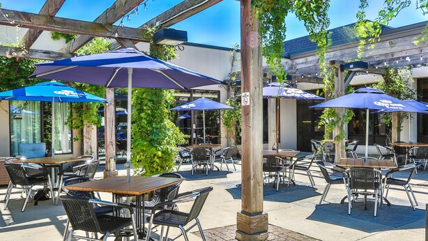 An outdoor patio area with tables and chairs, covered by a wooden pergola and blue umbrellas.