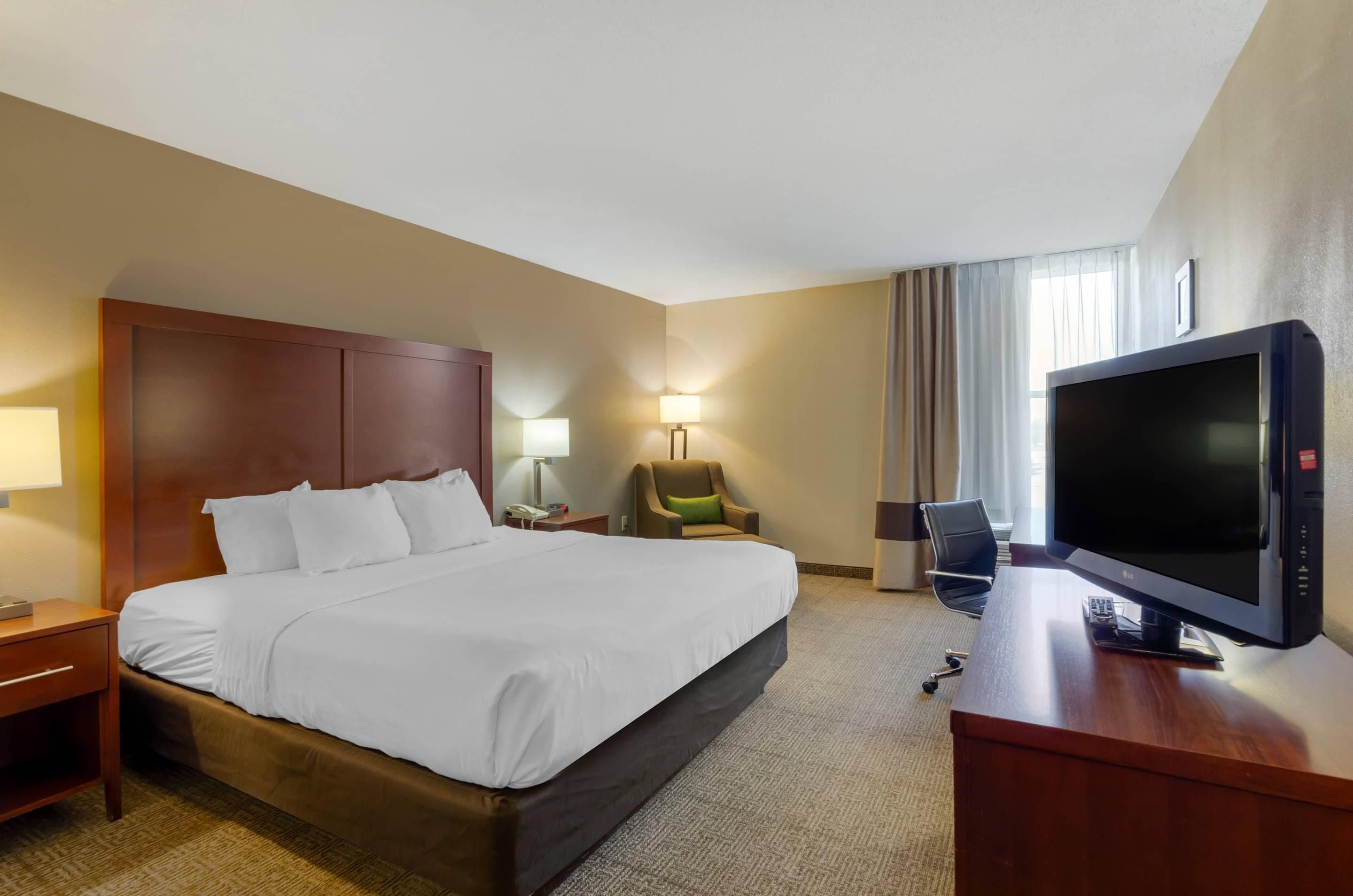 A hotel room with a large bed, a desk with a television, a chair, and a lamp.
