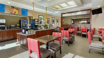 Restaurante de hotel con estación de café autoservicio, zona de estar con sillas rojas y mostrador con máquina de café.