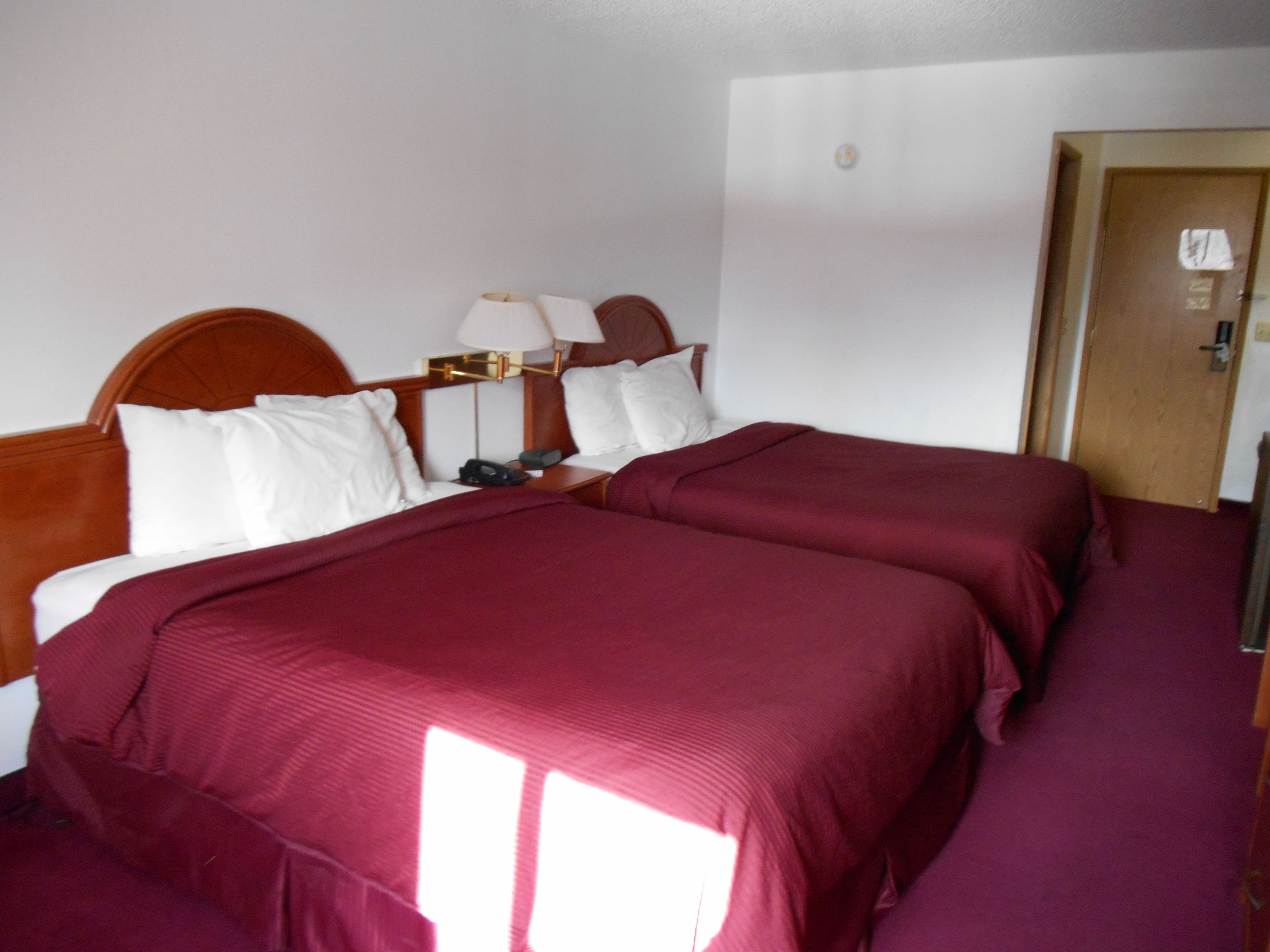 A hotel room with two beds, each with a wooden headboard, white pillows, and a red bedspread. There is a nightstand with a lamp and a phone.