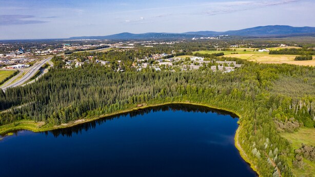 An aerial view of a lake surrounded by dense forest, with a small town and industrial buildings in the background. - Wedgewood Resort (Fairbanks)