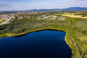 An aerial view of a lake surrounded by dense forest, with a small town and industrial buildings in the background. - Wedgewood Resort (Fairbanks)