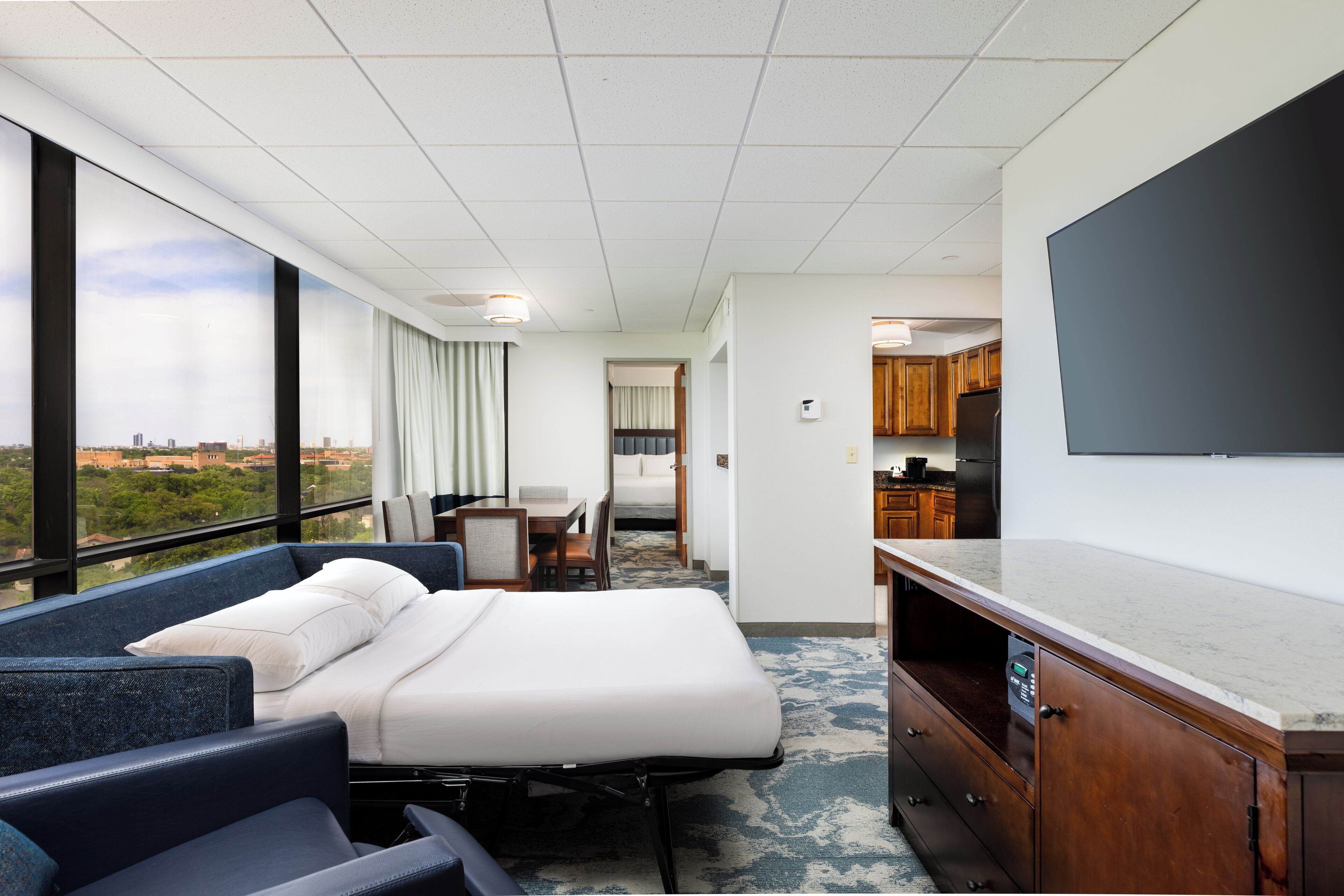 A hotel room with a bed, two chairs, a desk, a television, and a view of the city.