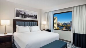 A hotel room with a large bed, two bedside tables with lamps, a bench, a cityscape view, and a framed picture on the wall. - DoubleTree by Hilton Houston Medical Center Hotel & Suites (Houston)