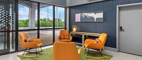Une pièce moderne avec de grandes fenêtres, des fauteuils à bascule orange, une petite table et un tableau encadré au mur.