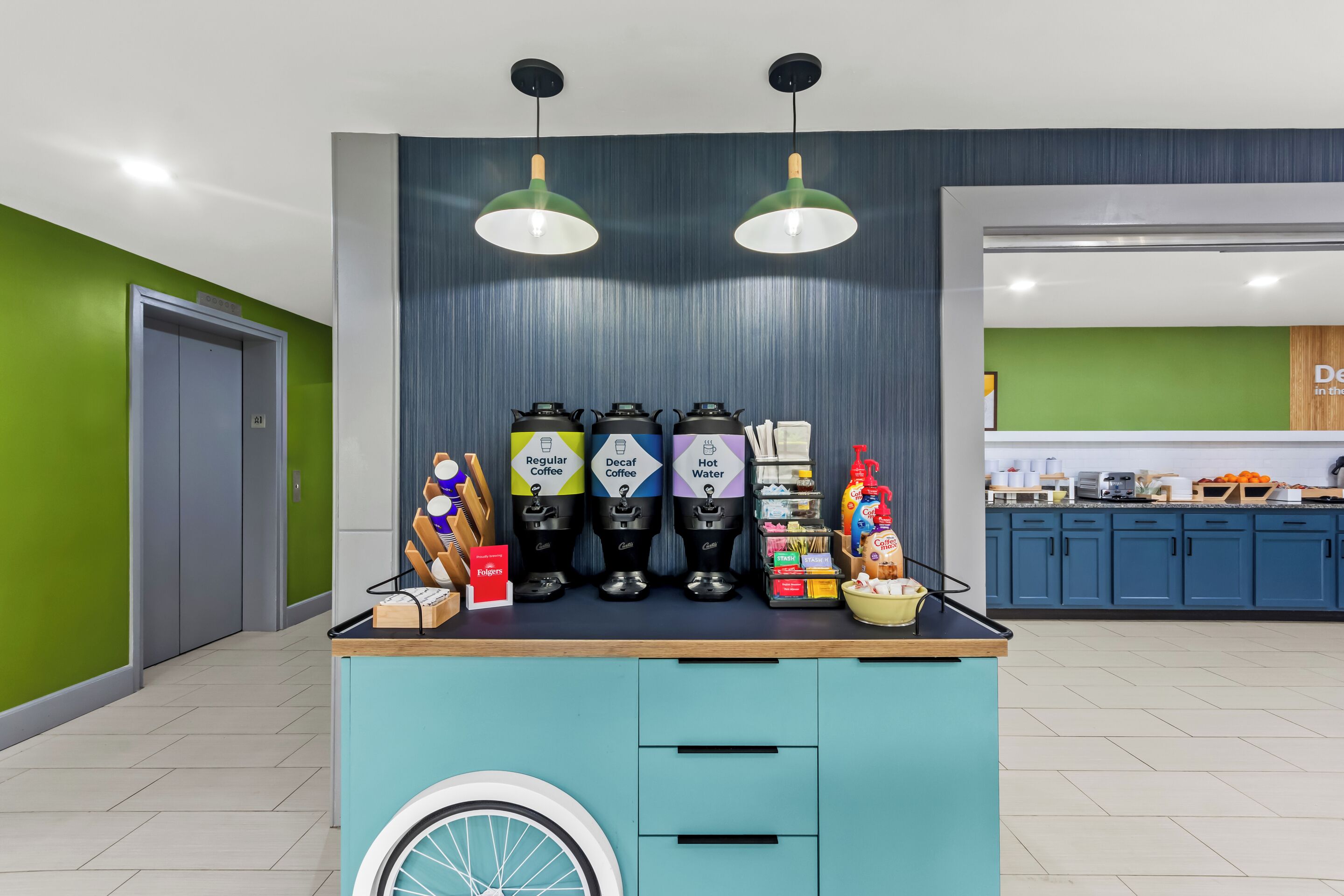 A coffee station with labeled dispensers for regular, decaf, and hot water, a bowl of fruit, and a tray of pastries.