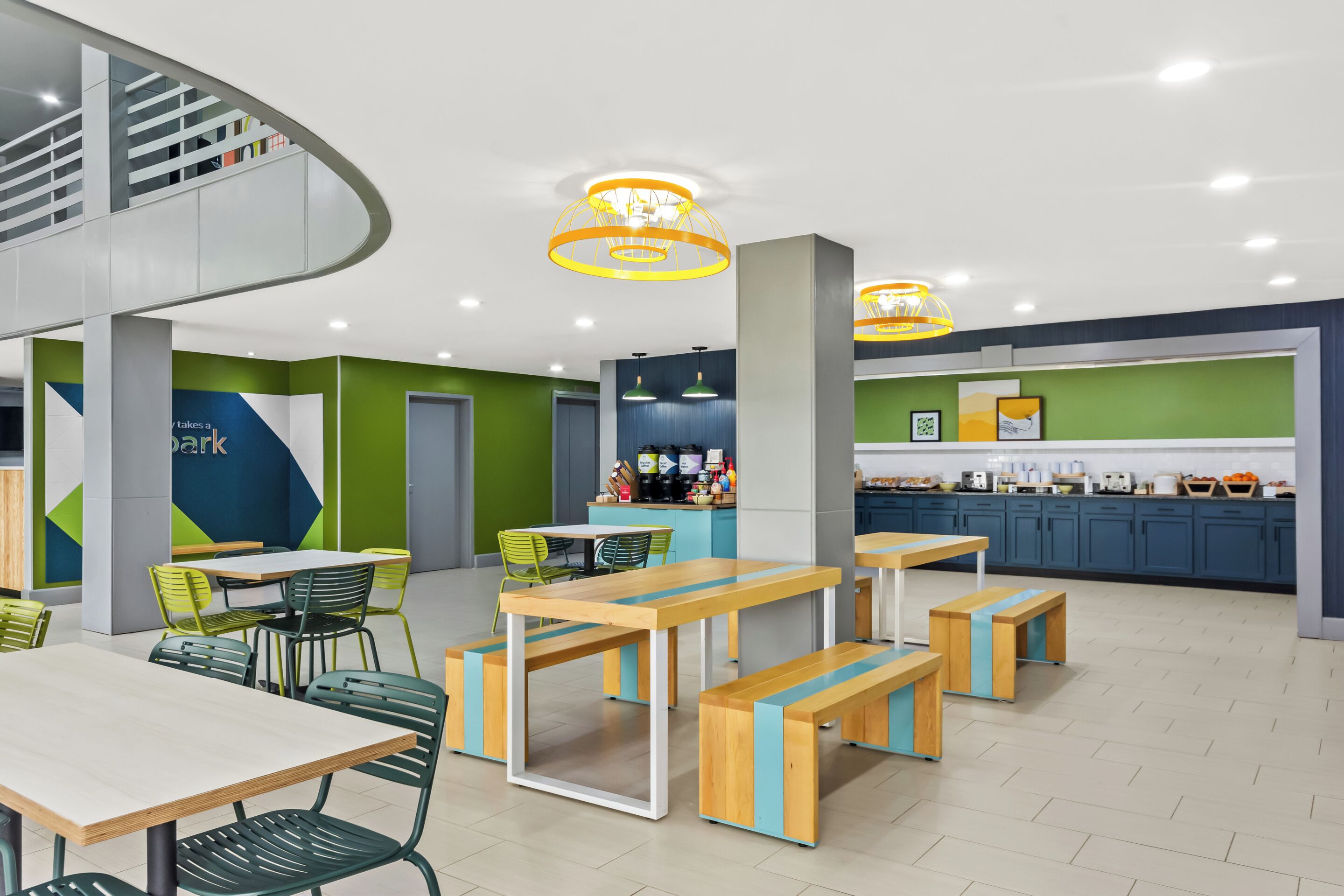 A modern indoor space with a curved staircase, wooden tables and benches, green chairs, and a kitchen area with blue cabinets.