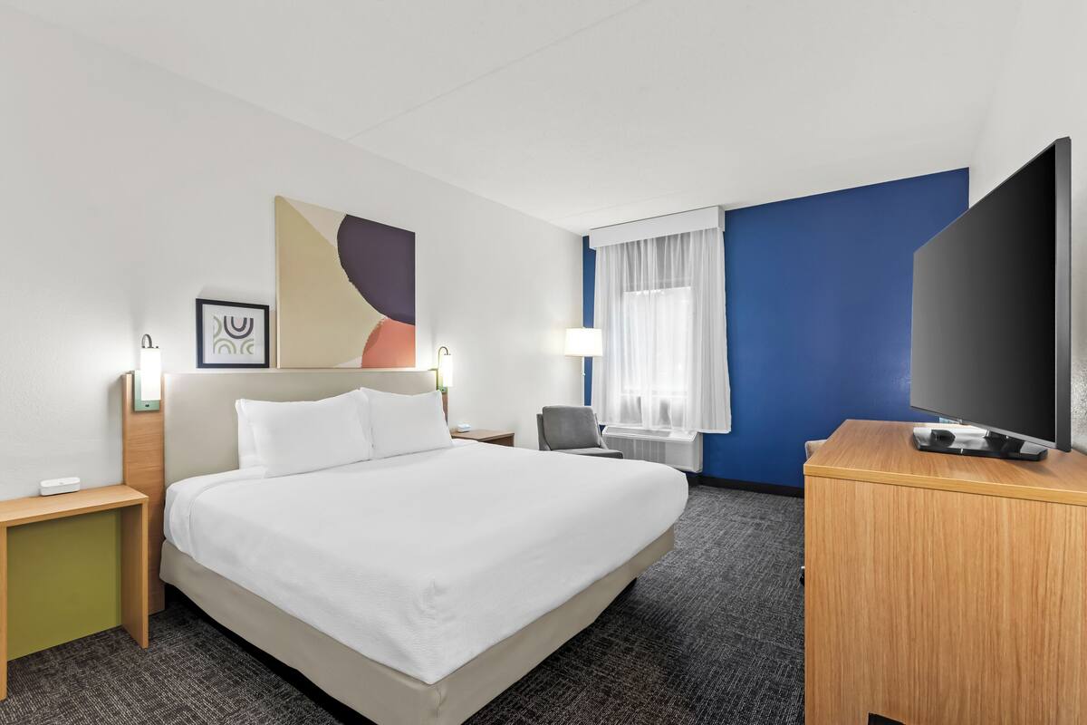 Ein Hotelzimmer mit einem großen Bett, einem Schreibtisch mit Flachbildfernseher, einem Sessel, einem Nachttisch und einer Wandlampe.