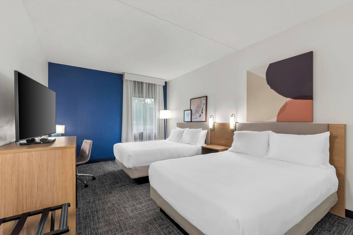 Ein Hotelzimmer mit zwei Betten, einem Schreibtisch, einem Fernseher und einem Fenster mit Vorhängen.