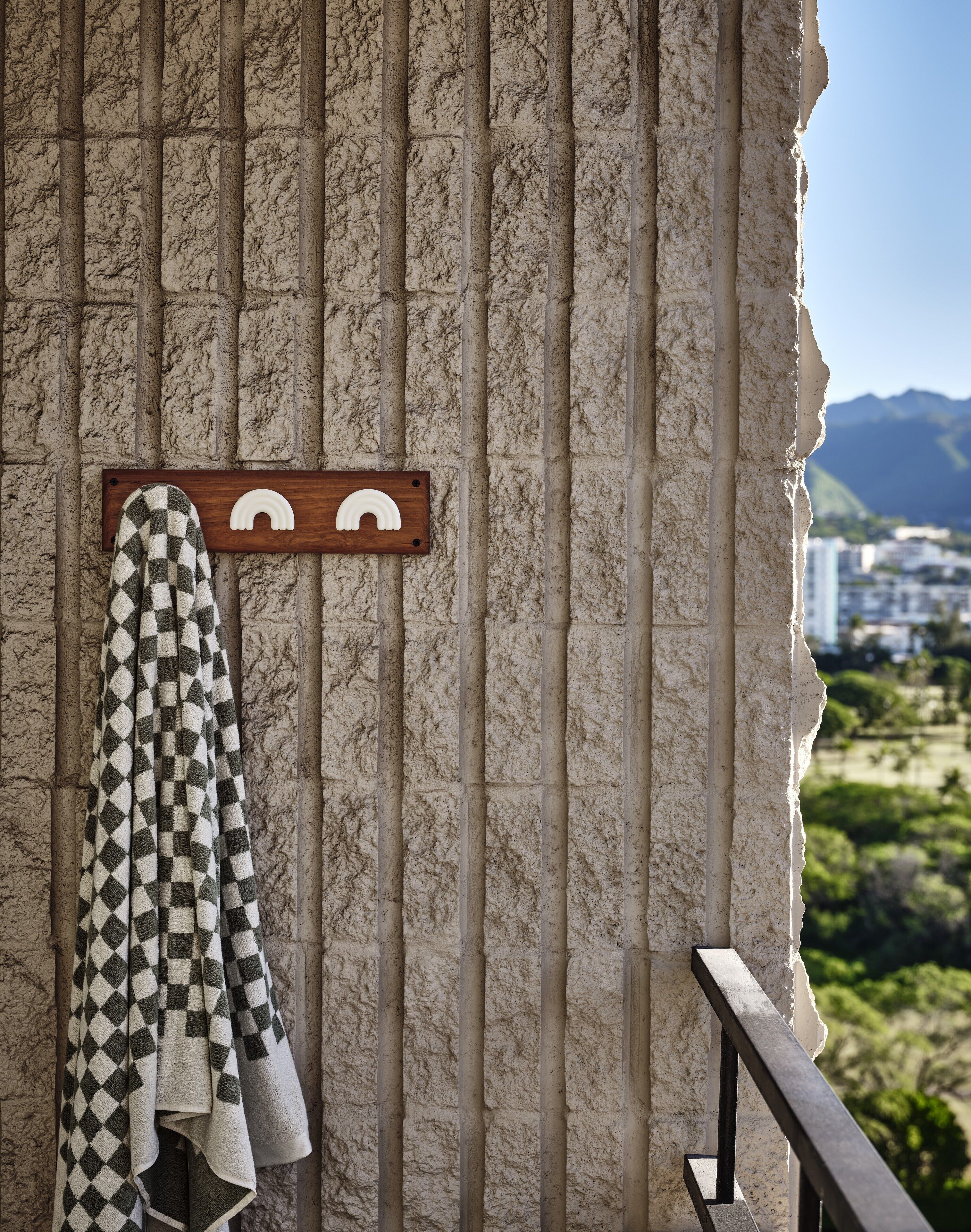 Un baño con una toalla de cuadros colgada en un perchero, un perchero de madera con una toalla y vistas a un paisaje urbano con montañas al fondo.