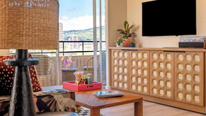 A modern living room with a large window, a flat-screen TV, a wooden cabinet with a woven design, and a wicker lamp.