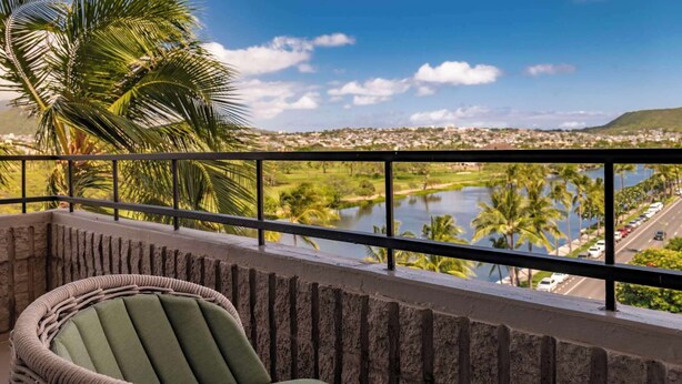 A balcony with a wicker chair, a view of a lake, palm trees, and a distant cityscape. - Wayfinder Waikiki (Honolulu)
