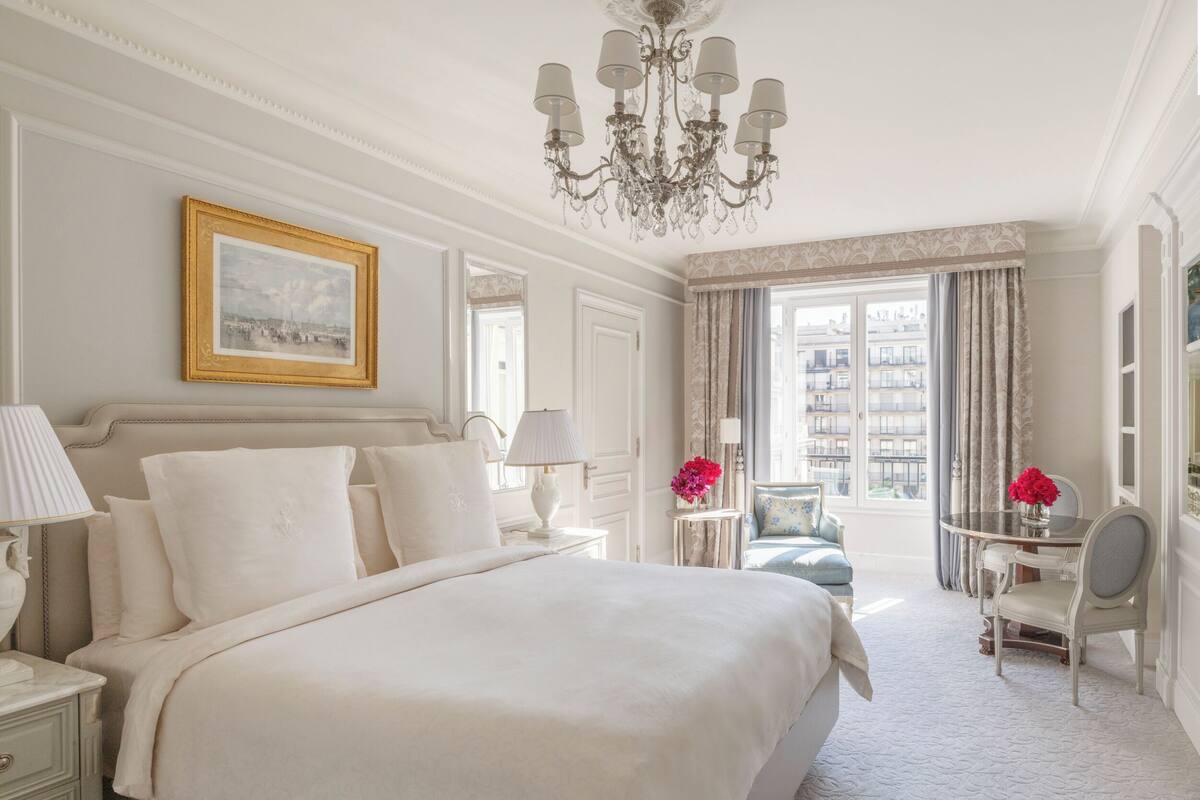 Une chambre spacieuse avec un grand lit, un lustre, un bureau avec une chaise et une vue sur la ville par la fenêtre.
