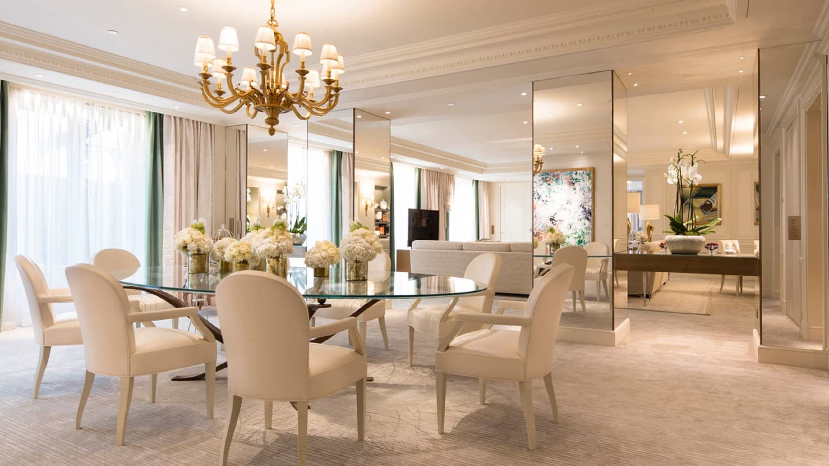 Une chambre d’hôtel spacieuse, dotée d’une table ronde en verre, de chaises blanches, d’un lustre et d’un grand miroir qui reflète la pièce.