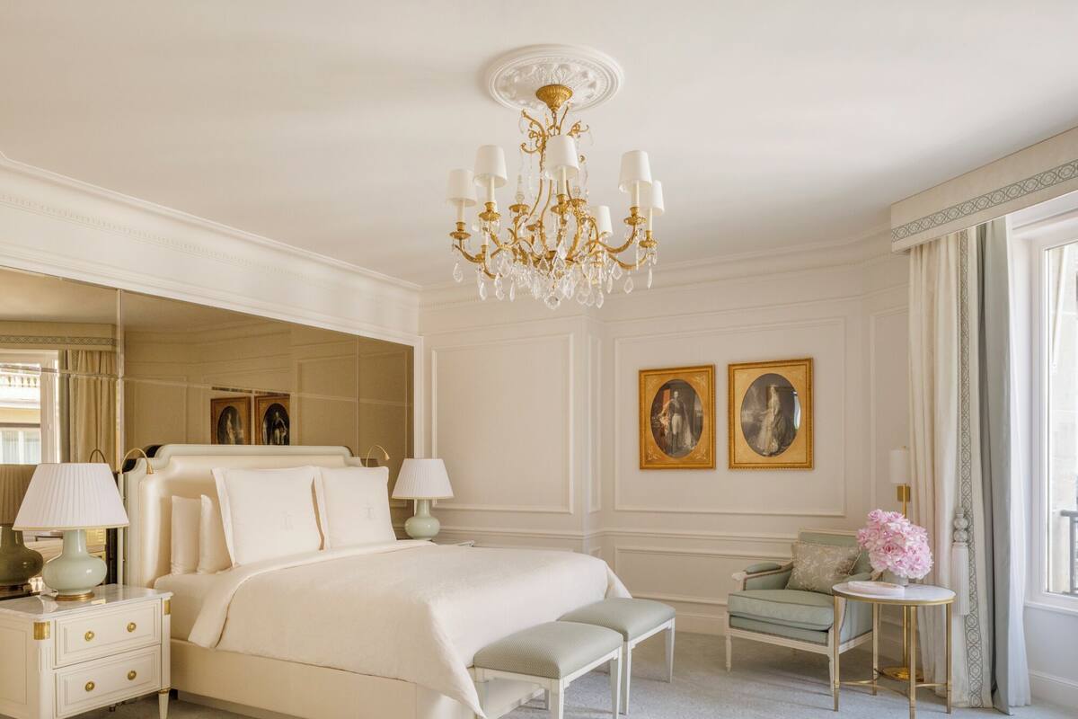 Une chambre spacieuse avec un grand lit, un lustre, un coin salon avec un fauteuil et une table, et deux tableaux encadrés au mur.