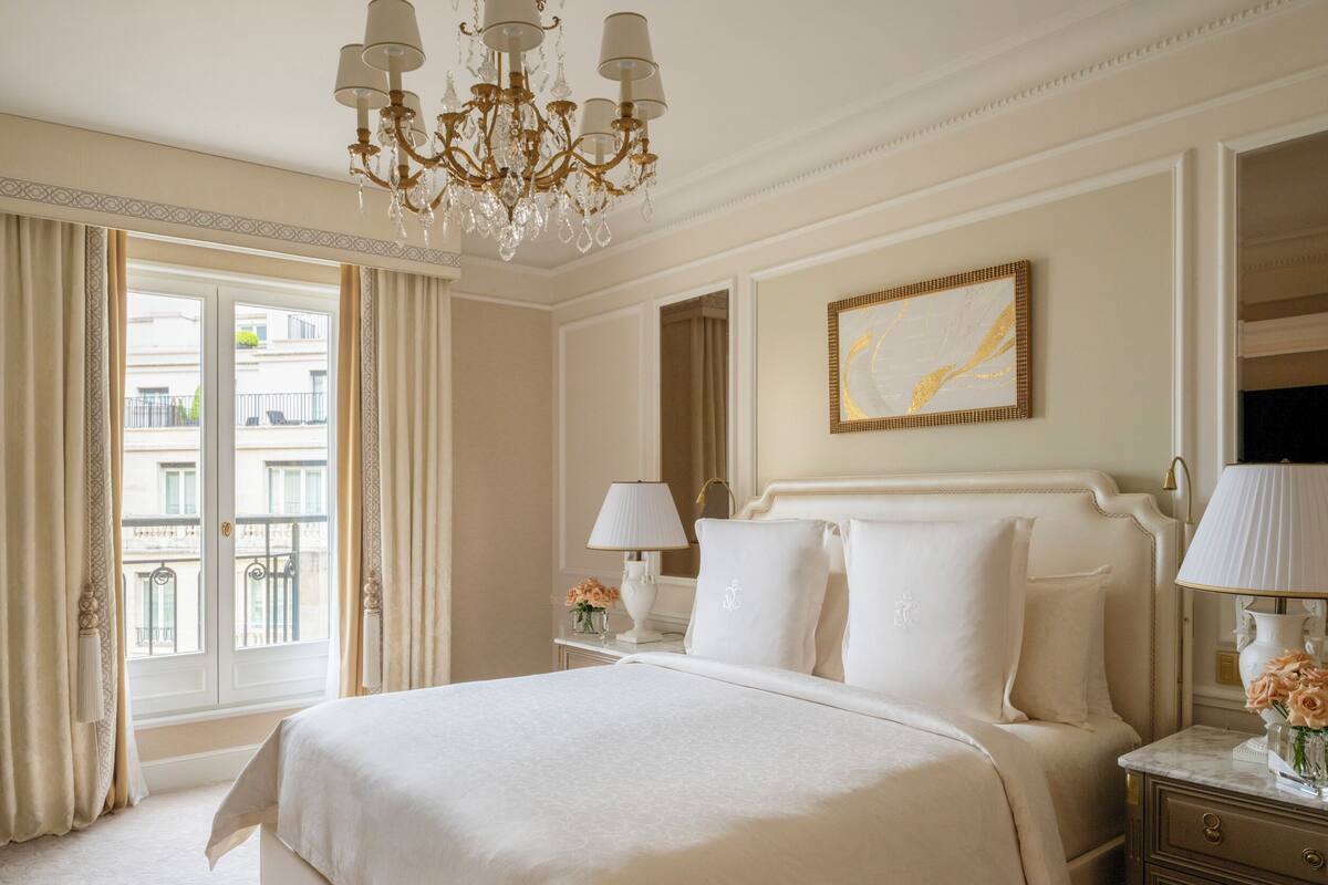 Une chambre spacieuse avec un grand lit, un lustre, un tableau et une vue sur la ville par la fenêtre.