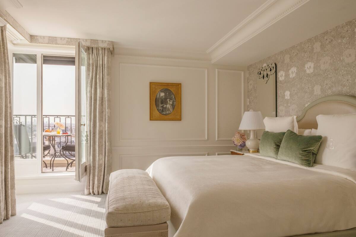 Une chambre d’hôtel avec un grand lit, une banquette, une table avec une chaise et un balcon avec des rideaux.